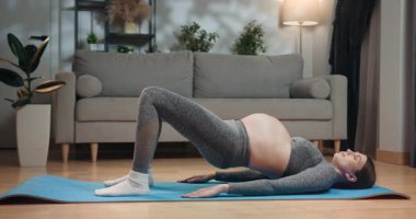 Aktif kıyafetli hamile kadın oturma odasında yoga minderinde briç pozu veriyor, hamilelik sırasında egzersiz yapıyor. Hamile Kadın Köprü Pozu Veriyor
