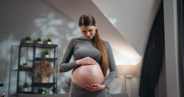 Genç bir kadın, sıcak bir yuva ortamında, yumuşak bir ifadeyle, nazik elleriyle hamile karnını kucaklar. Hamile Kadın Karnını Tutuyor