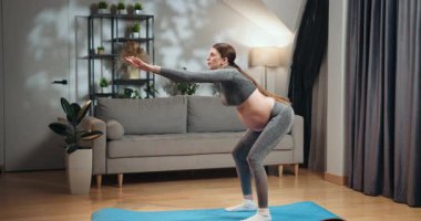 Gri aktif giysili hamile kadın oturma odasındaki koltuğu ve bitkileri olan mavi mindere çömeliyor. Hamile Kadın Evde Çömelme Yapıyor