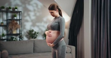 Atletizmde hamile bir kadın göbeğini takar, hafif ışıklı bir odada durur ve ev dekorları görünür olur. Hamile Kadın Karnını Tutuyor