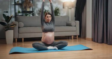 Hamile kadın nilüfer çiçeğinin üstünde yoga minderinde oturur, elleri karnına sarılır, oturma odasında farkındalık pratiği yapar. Hamile Kadın Evde Meditasyon Yapıyor