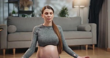 Rahat spor kıyafeti giyen hamile bir kadın yoga minderinde bacak bacak üstüne atmış, gözleri kapalı, rahat bir oturma odasında meditasyon yapıyor. Hamile Kadın Evde Meditasyon Yapıyor