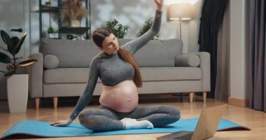 Gri aktif giysili hamile kadın oturma odasındaki mavi paspasın üzerinde yoga yapıyor. Yanında dizüstü bilgisayar var. Hamile Kadın Evde Yoga Yapıyor