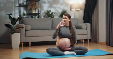 Yoga kıyafeti giymiş hamile bir kadın bacak bacak üstüne atarak mindere oturur, içme suyu içer, karnını tutar, evde kanepe ve bitkiyle birlikte. Su Yogası İçen Hamile Kadın