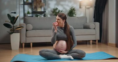 Hamile kadın yoga minderinde bağdaş kurmuş, elinde elmayla oturuyor, gri atletik giysiler giyiyor, kapalı bir yerde kanepe ve arka planda bitkiler var. Mat 'te Elma Yiyen Hamile Kadın