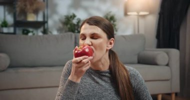 Gri spor giysili hamile bir kadın evde kırmızı elma yiyor ve hamileliği boyunca sağlıklı beslenmeye odaklanıyor. Elma Yiyen Hamile Kadın