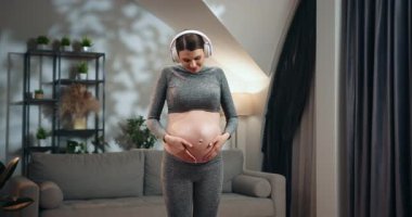 Hamile bir kadın kulaklık takıyor ve oturma odasında durup karnına dokunuyor. Hamilelik boyunca müzik ve rahatlamanın tadını çıkarıyor. Hamile Kadın Müzik Dinliyor