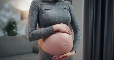 Gri atletik giysili hamile kadın iki eliyle yuvarlak göbeğini, arka planda yumuşak ışık ve perdelerle kapalı bir yerde giyiyor. Hamile Kadın Karnını Tutuyor