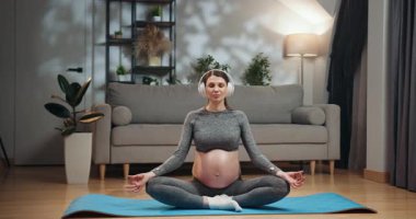 Spor kıyafetli hamile bir kadın yoga minderinde kulaklık takarak oturur ve oturma odasında gözleri kapalı meditasyon yapar. Hamile Kadın Kulaklıkla Meditasyon Yapıyor