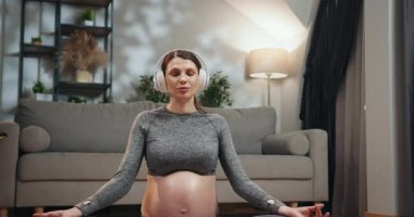 Spor giyim bölümünde hamile bir kadın kapalı gözlerle meditasyon yapıyor. Kapalı yerde yoga minderinde kulaklık takıyor. Hamile Kadın Kulaklıkla Meditasyon Yapıyor