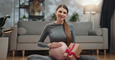 Bekleyen anne kucağında hediye kutusuyla yerde oturur. Oturma odasında kanepe ve bitkiyle. Hediye Kutusu Olan Hamile Kadın