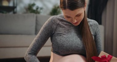 Gri elbiseli hamile kadın, elinde kırmızı kurdeleyle bağlı kahverengi bir hediye kutusu tutarken göbeğini sallıyor. Hamile Kadın Kulaklıkla Meditasyon Yapıyor