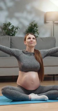 Gri aktif giysili hamile kadın mavi yoga minderi üzerinde meditasyon yapıyor. Elleri dua pozisyonunda birbirine bastırılmış. Gözleri kapalı.