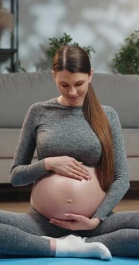 Gri aktif giysili hamile bir kadın yoga minderinde oturuyor. Sevgi dolu bir ifadeyle karnına hafifçe dokunuyor.
