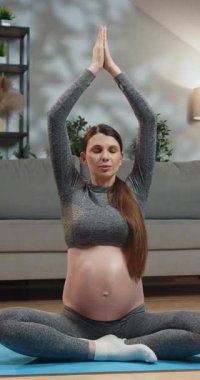 Rahat spor kıyafeti giyen hamile bir kadın yoga minderinde bacak bacak üstüne atmış, elleri başının üstünde, gözleri huzurlu meditasyonda kapalı oturuyor.