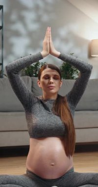 Gri aktif giysili hamile kadın elleri bağdaş kurup dua pozisyonunda oturuyor, gözleri kapalı, oturma odasında huzur içinde meditasyon yapıyor.