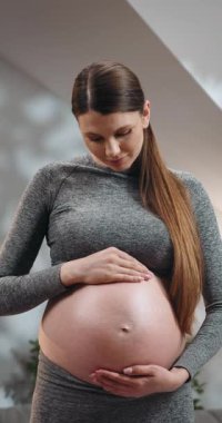 Genç bir kadın hamile karnını nazikçe eğiyor, sevgi dolu bir ifadeyle aşağıya bakıyor, anneliği bekliyor.