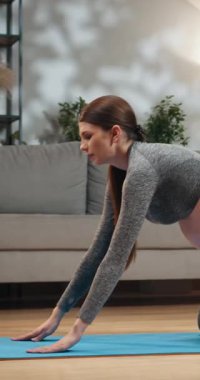 Gri spor giysili genç bir kadın oturma odasında yoga yapıyor. Arka planda bir raf ve kanepe var.