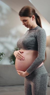 Sporcu olan genç bir kadın hamile karnını sarkıtır ve kameraya doğru nazikçe bakar.