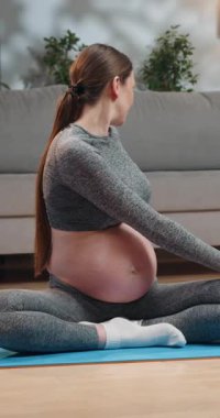 Atletik giysili hamile kadın oturma odasındaki mavi paspasın üzerinde omurga bükme yoga pozu veriyor.