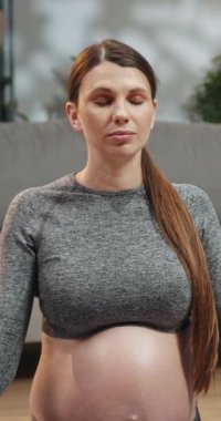 Spor giyim sektöründe hamile bir kadın oturma odasında yoga minderinin üzerine gözlerini kapatarak meditasyon yapıyor. Refah için farkındalık ve rahatlama egzersizi yapıyor.