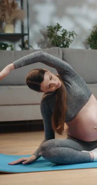 Gri aktif giysili hamile kadın oturma odasında mavi yoga minderine uzanıyor, internetteki dersler için dizüstü bilgisayarı yakınlarda.