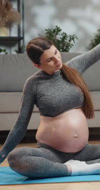 Gri aktif giysili hamile kadın içeride mavi yoga minderini eğip doğum öncesi sağlık ve refahına odaklanıyor.