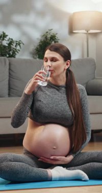 Hamile bir kadın yoga minderinde bağdaş kurmuş oturur. Camdan, evden, rahat atmosferden su içer.