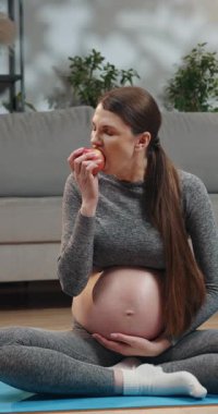Spor giyimli hamile bir kadın yoga minderinde bacak bacak üstüne atarak oturur. Elma yer, kapalı yer, sağlıklı yaşam tarzı, üçüncü üç aylık dönem.