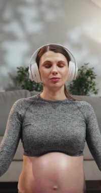 Spor kıyafetli hamile bir kadın yoga minderinde kulaklık takarak oturur ve gözleri kapalı meditasyon yapar.