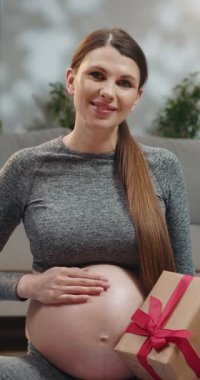 Bekleyen anne, elinde kırmızı kurdeleyle küçük bir hediye kutusu tutarken karnını kucaklar.