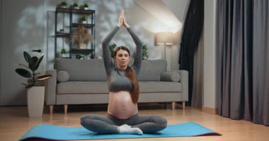 Gri aktif giysili hamile kadın yoga minderinin üzerinde nilüfer pozisyonunda oturuyor, elleri kapalı, gözleri kapalı, evde farkındalık pratiği yapıyor. Hamile Kadın Evde Meditasyon Yapıyor
