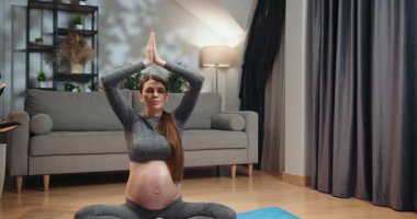 Spor kıyafetli hamile bir kadın yoga minderinde bacak bacak üstüne atarak oturur, dua pozunda ellerini birbirine bastırarak meditasyon yapar, gözleri kapalı, rahat bir oturma odasında. Hamile Kadın Evde Meditasyon Yapıyor