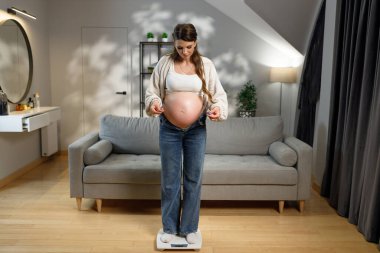 Bekleyen anne oturma odasında tartıda duruyor, hamilelik sırasında kilo alımını kontrol ediyor, kıyafetleri sıkıyor.