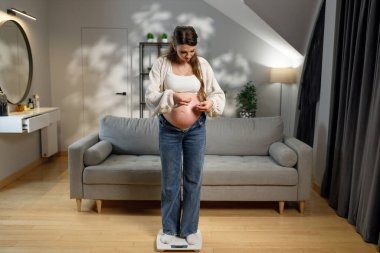 Hamile kadın, karnını kontrol ederek, hamilelik yolculuğunu ve evdeki vücut değişikliklerini sembolize ediyor.