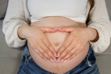 Kadınların elleri hamile karnı üzerinde kalp şekli oluşturur, yakın plan görüntüsü, yeni bir hayat için sevgi ve beklenti taşır.