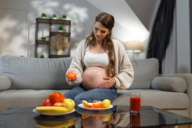 Bekleyen anne kanepeye oturur, elinde hurma dilimi, masada sağlıklı yiyecekler, ev ortamları, sıcak ışıklandırma.