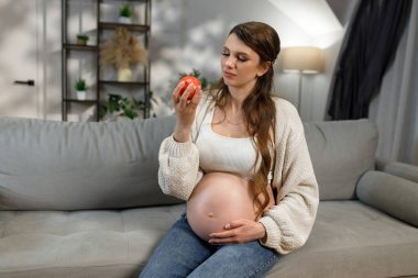 Hamile kadın, elinde kırmızı elmayla kanepede oturur ve hamilelik boyunca sağlıklı beslenmeyi düşünür.