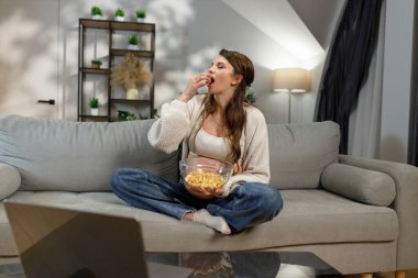 Genç bir kadın kanepede bacak bacak üstüne atarak oturuyor, dizüstü bilgisayardan bir şeyler izlerken kaseden patlamış mısır yiyor.