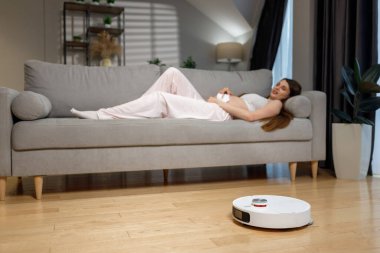 Hamile kadın kanepede dinlenirken robotik elektrikli süpürge oturma odasındaki ahşap zeminde çalışıyor.