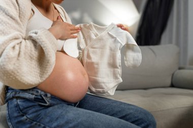 Anneler kanepeye oturup karnına beyaz bebek tulumu dayayarak yeni bir hayat bekliyor ve bebek eşyaları hazırlıyor.