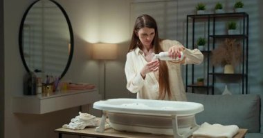 Kadın, su ve baloncuklarla dolu taşınabilir bebek küvetine dikkatlice sıvı sabun ekliyor. Anne Bebek Banyosu Hazırlıyor