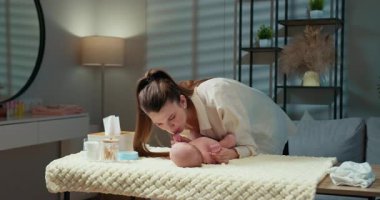 Anne, bebek ayaklarını yumuşak bir battaniyede, rahat bir odada bebek bezi değiştirirken nazikçe öper. Anne Öpüşen Bebek Ayağı