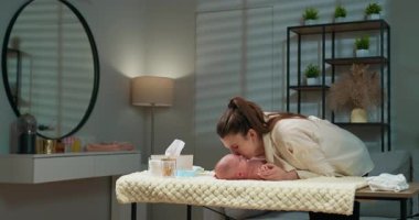 Anne yeni doğmuş bebeğini nazikçe masasını değiştirirken öpüyor, sıcak bir bebek odasında sevgisini ve bakımını ifade ediyor. Anne Yenidoğan Bebeği Şefkatle Öpüyor