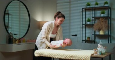 Anne, yeni doğmuş bebeğini rahat, modern bir bebek odasında, soyunma masasında nazikçe giydirir.
