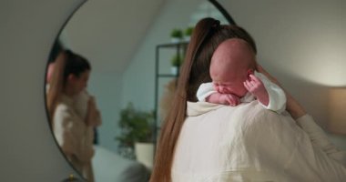 Anne, yeni doğmuş bebeğini, sahneyi yansıtan bir aynayla hafifçe aydınlatılmış bir odada omzunun üzerinde tutuyor. Anne kucağında yeni doğan bebek