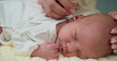 Anne, yeni doğmuş bebeklerin kulaklarını pamuklu bezle nazikçe temizlerken bebek de yumuşak battaniyenin üzerinde huzur içinde uyuyor. Bebek Kulak Temizleme Bakımı