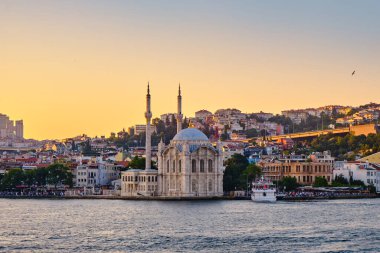 Boğazdaki ortakoy iskelesi, gün batımında şehrin arka planındaki caminin manzarası. Feribotlar rıhtımda duruyor, insanlar iskelede yürüyor.