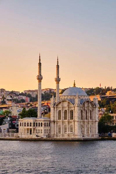 Boğazdaki ortakoy iskelesi, gün batımında şehrin arka planındaki caminin manzarası. Feribotlar rıhtımda duruyor, insanlar iskelede yürüyor.