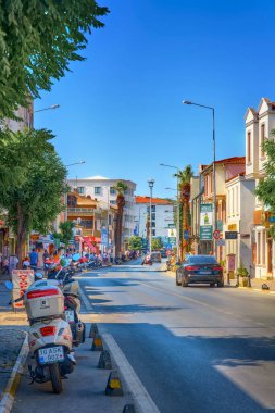 Ayvalik, Türkiye, 12 Temmuz 2025. Central City caddesinde park etmiş motosikletler, arabalar, yerel dükkanlar, palmiye ağaçları ve açık mavi gökyüzü altında yayalar. Popüler bir Ege bölgesinde yerel yaşam tarzı, ulaşım ve mimariyi yansıtan şehir manzarası